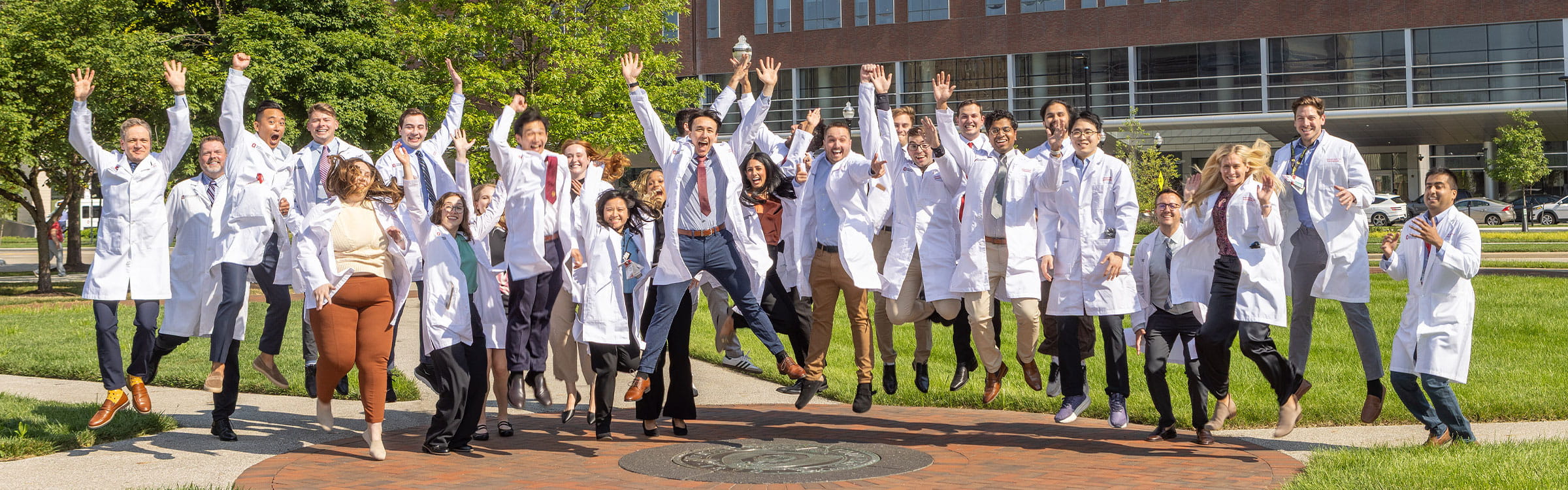 A group of smiling doctors dressed in white coats energetically jumping on the OSU seal in front of The James Cancer Hospital and the New University Hospital.