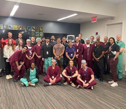 Large group of people huddled together wearing scrubs in front of an anesthesia machine and a balloon bunch next to them .