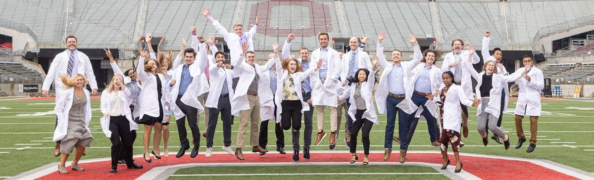 Residents wearing white labcoats jumping all together at once at the OSU Football Stadium 50 yard line.