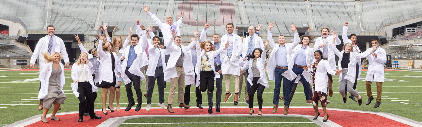 Residents wearing white labcoats jumping all together at once at the OSU Football Stadium 50  yard line.
