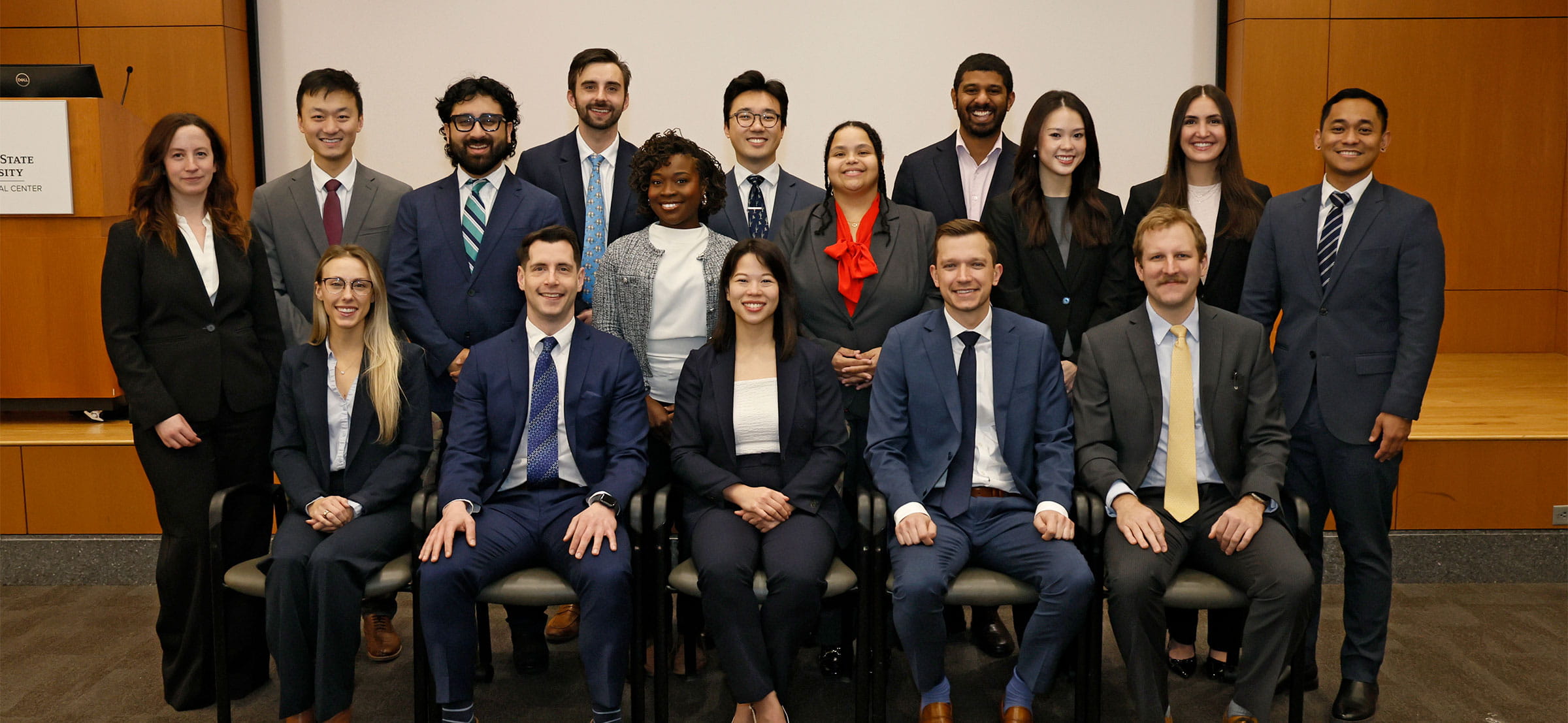 A diverse group of 16 professionals in formal attire pose together, smiling in a conference room setting, conveying unity and professionalism.