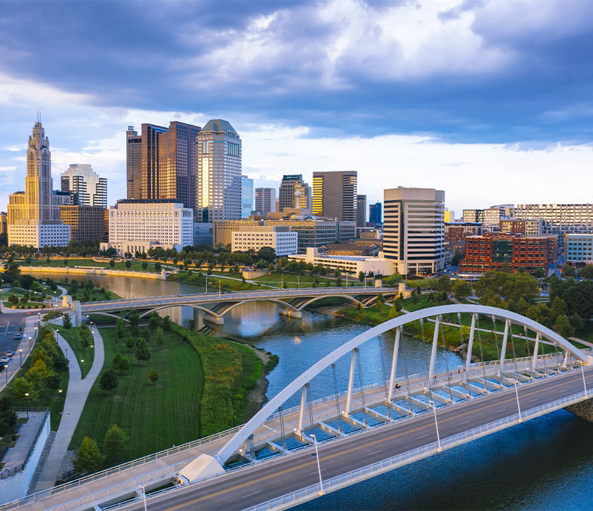 Aerial view of Columbus skyline under a blue sky with clouds. Tall buildings reflect sunlight, a river flows through, and bridges and greenery are visible.
