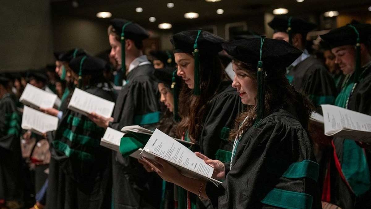 College of Medicine Spring 2022 graduating class reading from booklet wearing graduation robes in auditorium