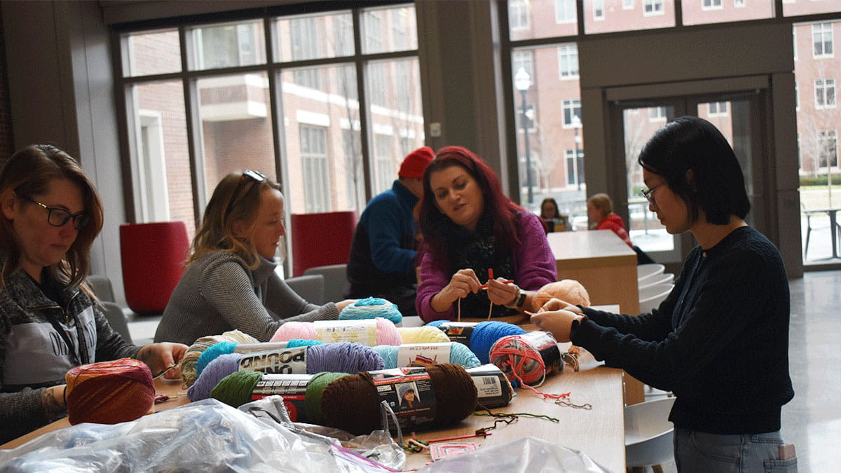 A group of four people sitting around a table in a bright room, focused on knitting.