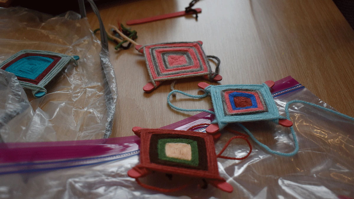 Colorful woven crafts, featuring square patterns with sticks at each corner, rest on a wooden table near plastic bags.