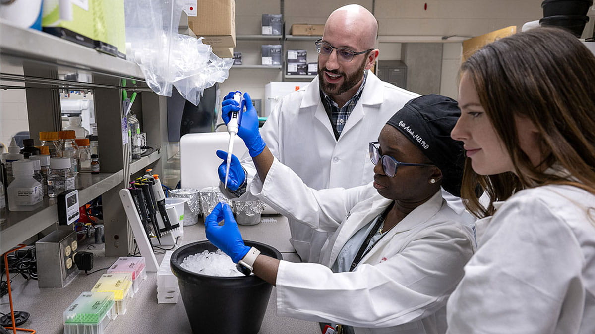 Three scientists in lab coats work together in a laboratory. One uses a pipette, while the others observe.