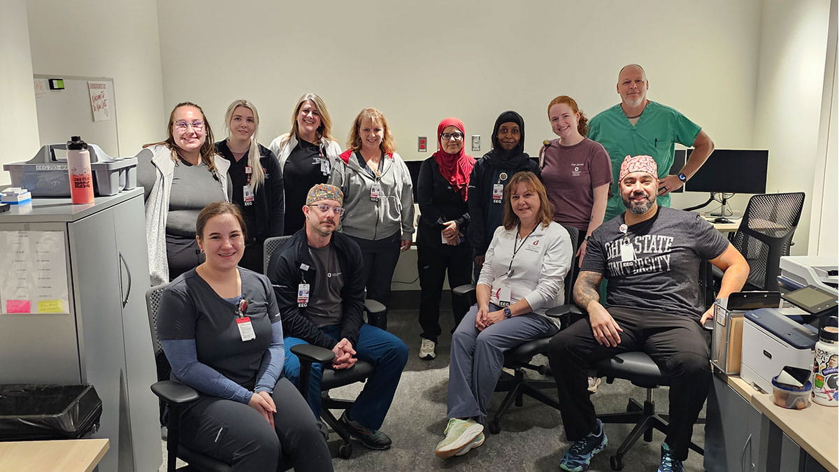 A diverse group of healthcare workers, wearing badges and casual attire, pose smiling in an office setting, conveying teamwork and camaraderie.