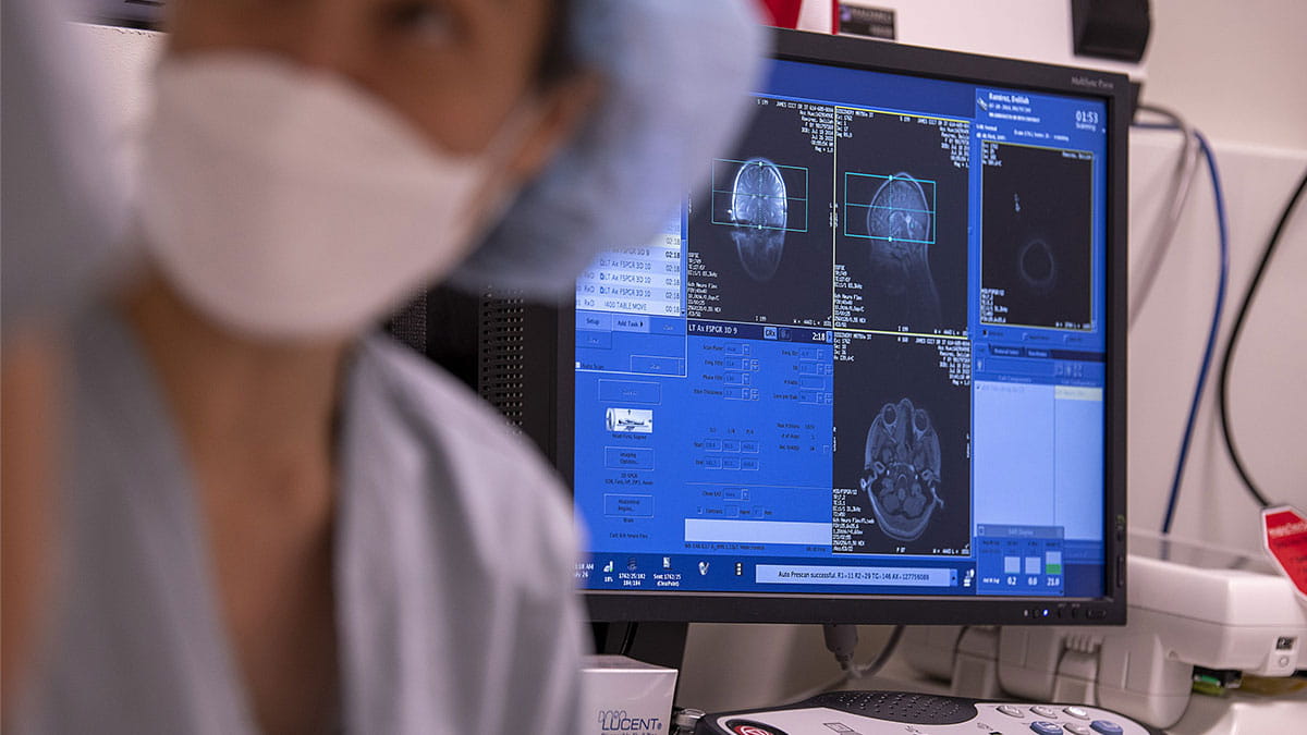 The image features a hospital or clinic setting. In the foreground, a medical professional wearing a mask and scrubs is partially visible and out of focus, creating a sense of depth. The main focus is on a computer monitor displaying several MRI images. These images show cross-sectional views of a human brain