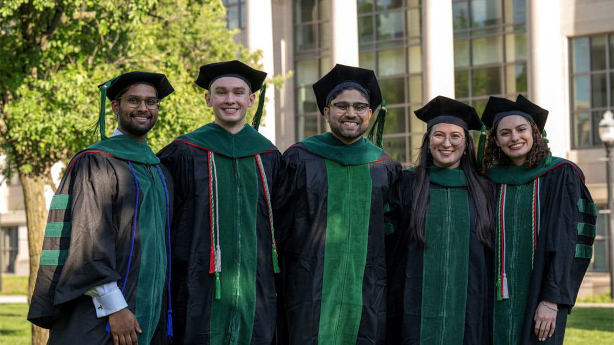 Five graduates in black and green gowns and caps smile outdoors in front of a building with columns, conveying joy and accomplishment.