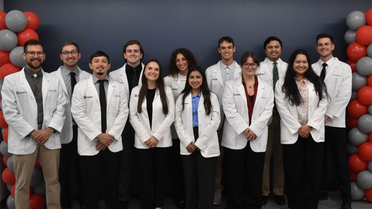 A group of people in white coats stand together next to a balloon arch.