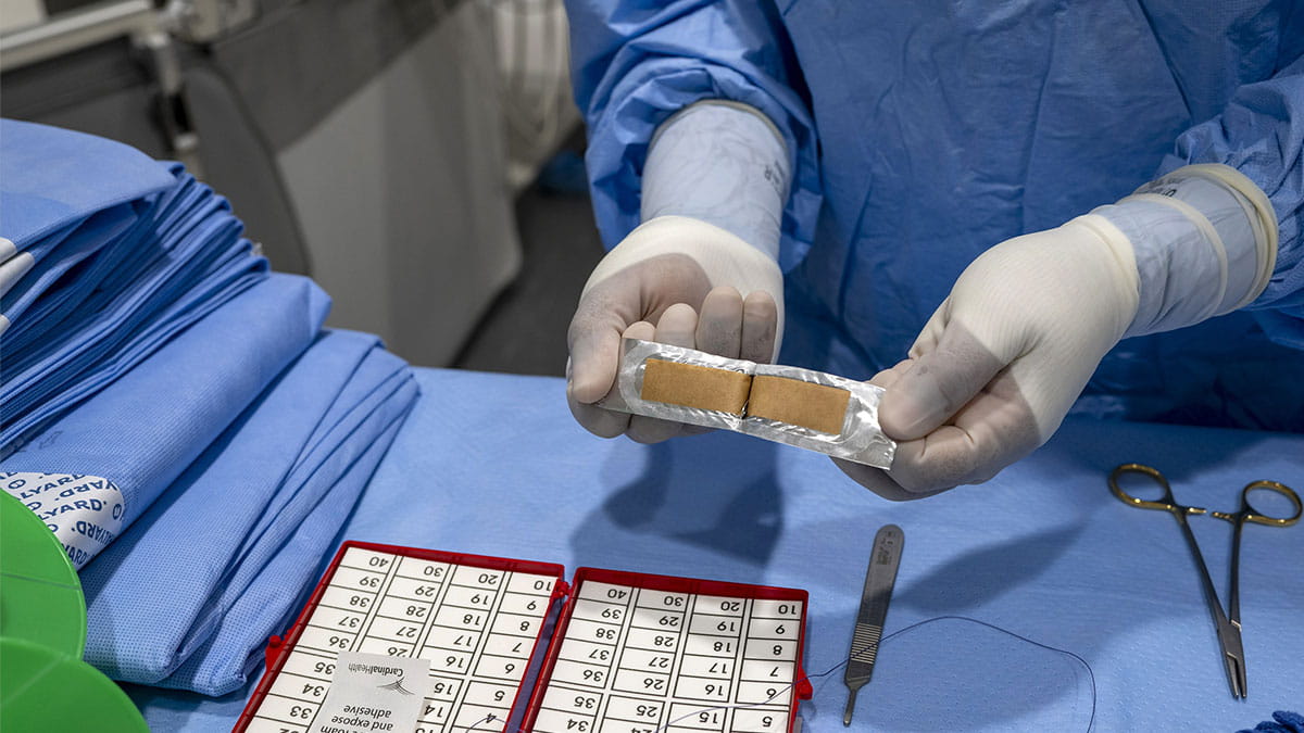 A medical professional in blue scrubs and gloves holds a sterile package with two brown bandages, with a surgical count sheet and instruments on a blue drape nearby.