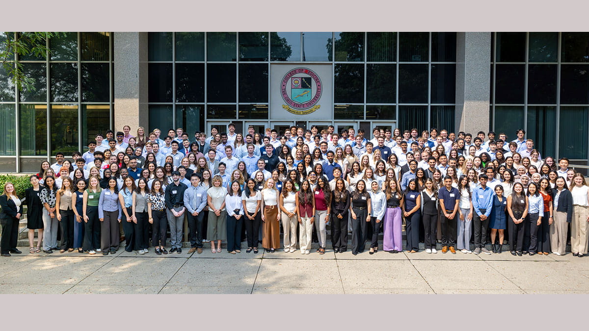 A large group photo of students in business attire standing in front of a glass building, with diverse individuals and a university seal on display.