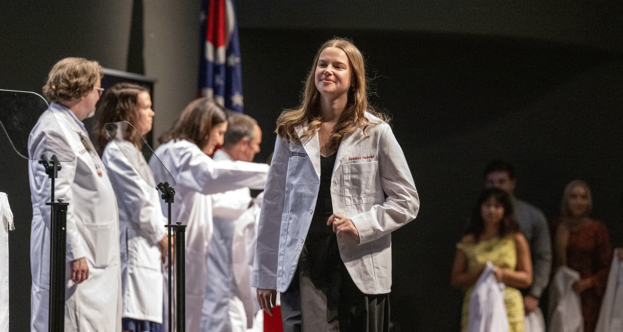 A young woman in a white coat smiles as she walks on stage during a coat ceremony, surrounded by others in white coats and a seated audience.