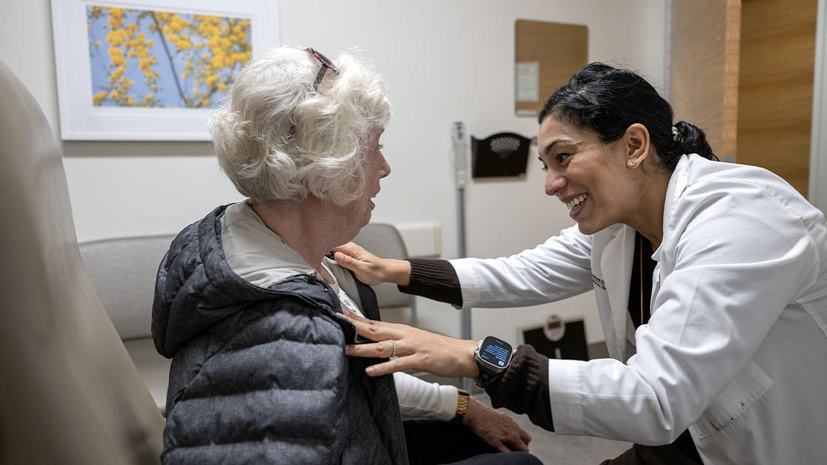 A doctor in a white coat smiles while gently touching the shoulder of an elderly female patient in a medical office.