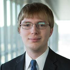 Patrick Stevens is wearing a black suit with a blue tie. He is smiling slightly, has blonde hair with a bowl cut, and is wearing glasses reflecting something green in the distance. He is standing on a brightly lit glass windowed bridge. 