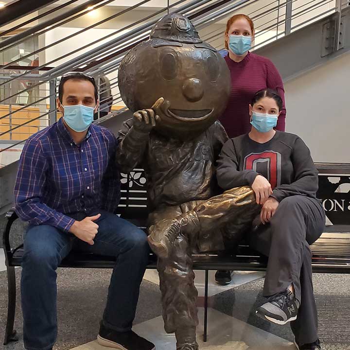 Three team members wearing masks sitting on bench with statue of Brutus Buckeye
