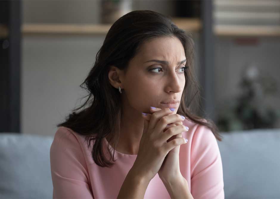 Woman sitting on couch inside with hands folded looking concerned