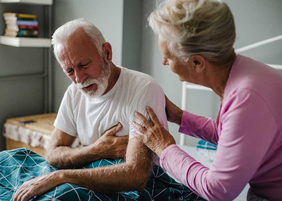 Senior man suffering from chest pain at home with senior woman checking on him